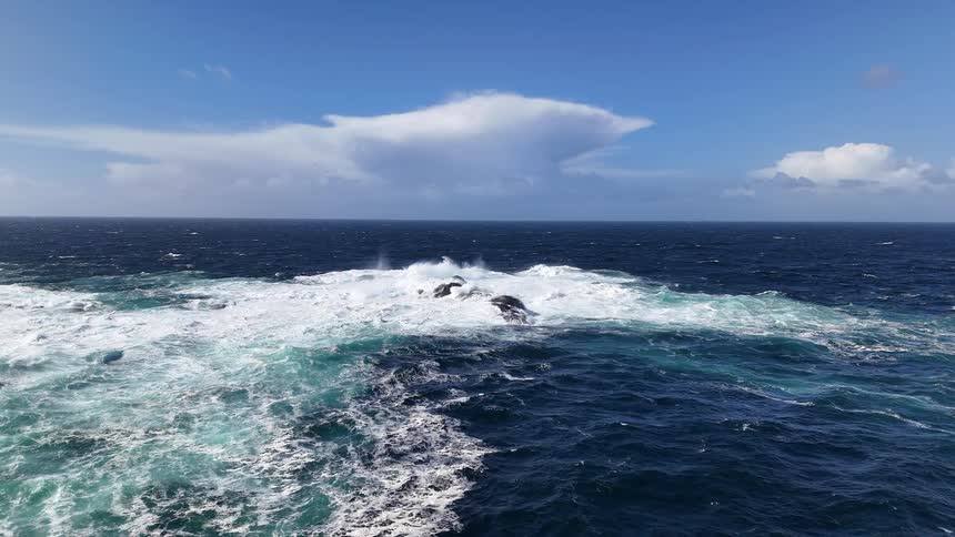A super slow motion aerial view of powerful ocean waves crashing against rugged rocks on a beautiful sunny day with blue skies
