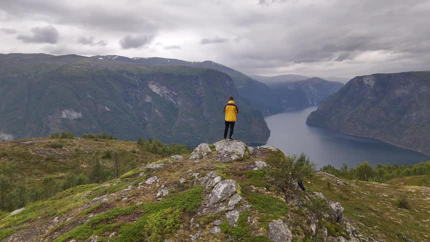 Slow aerial pull-back reveals a hiker in a yellow jacket standing on a mountain peak overlooking the vast and stunning Aurlandsfjord in Norway