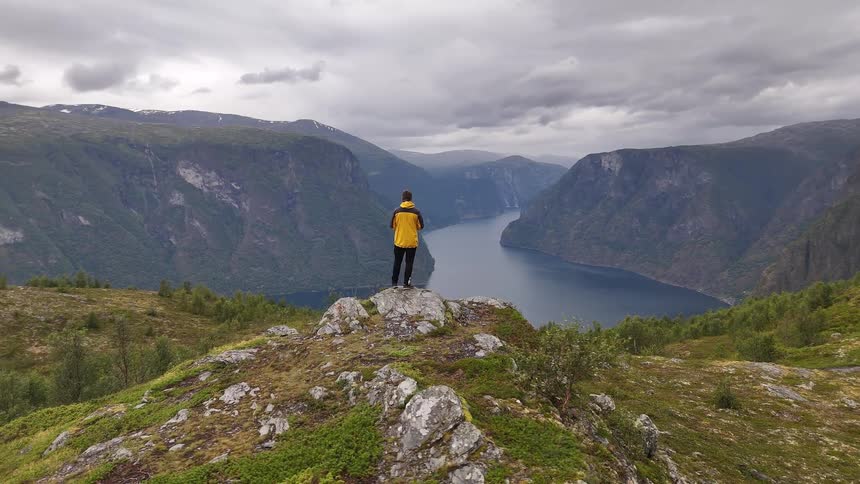 An adventurous man in a yellow jacket stands on a mountain peak, soaking in the epic view of a vast fjord on a cloudy day