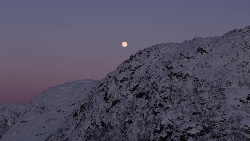 A timelapse captures the full moon rising from behind a snowy mountain peak against a purple and pink twilight sky