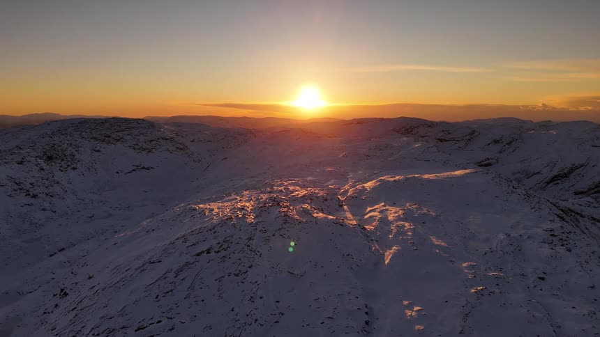 A static aerial view shows a vast, snow-covered mountain range illuminated by a golden sunset on the horizon