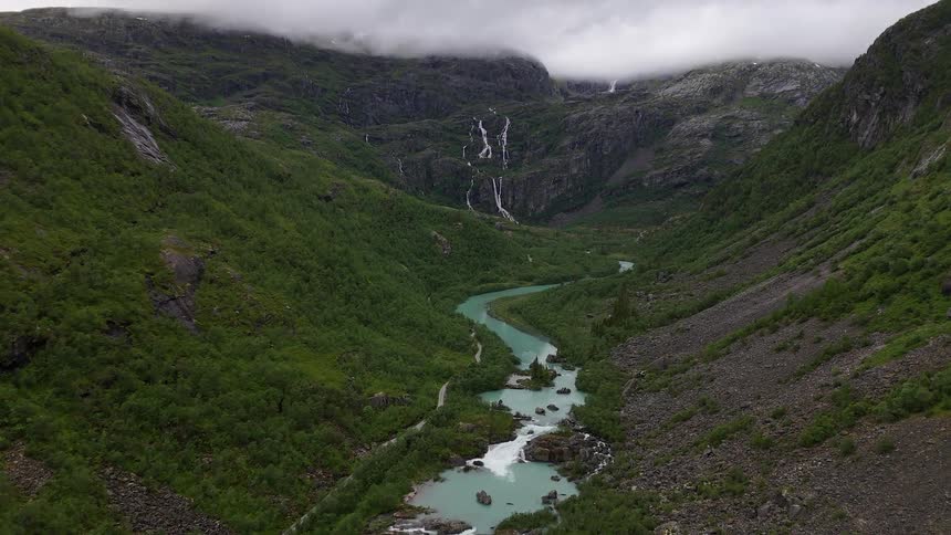 A slow aerial flight through a lush green valley reveals a winding turquoise glacial river and cascading waterfalls under a cloudy sky