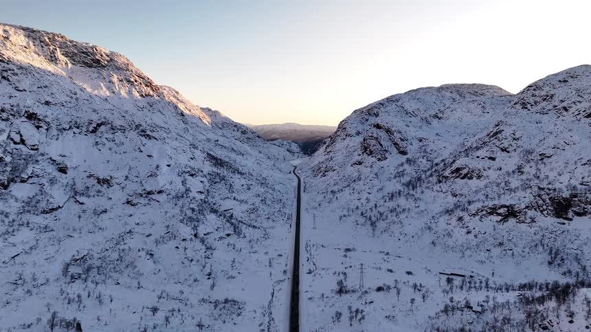 An aerial view of a single road stretching through a vast, snow-covered mountain valley at sunrise