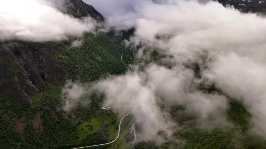 High-angle aerial view of a lush green mountain valley with a winding road, partially obscured by beautiful white clouds