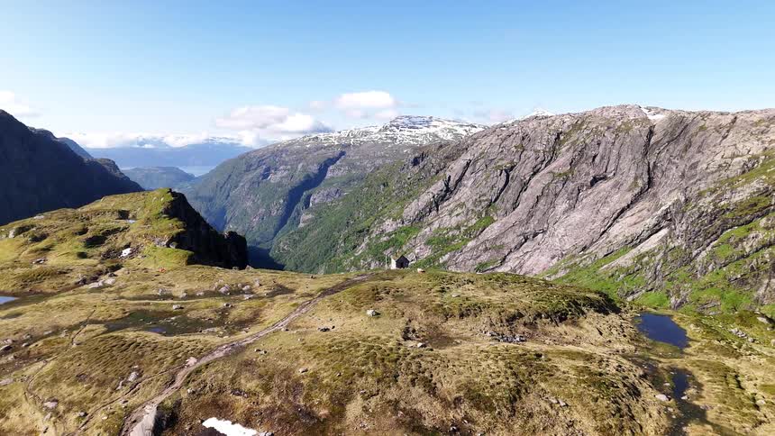 An epic aerial pullback from a small mountain hut reveals a stunning, sun-drenched valley and fjord in Norway