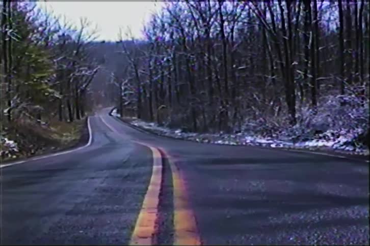 A static shot of a lonely, winding country road through a snow-dusted forest with a vintage VHS aesthetic