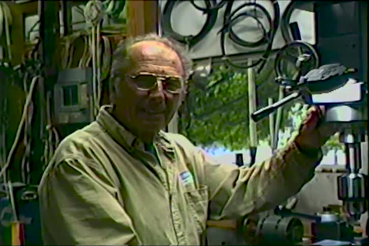 An elderly man with glasses pauses from working on machinery in his workshop to smile at the camera