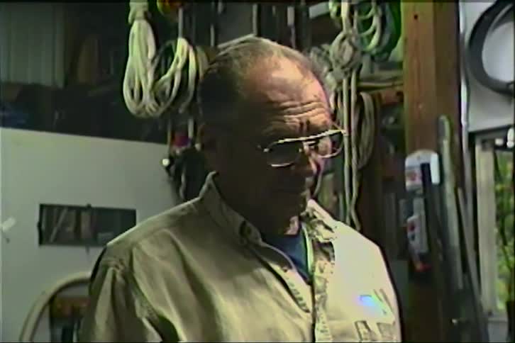 A vintage VHS portrait of an elderly craftsman standing thoughtfully in his cluttered workshop