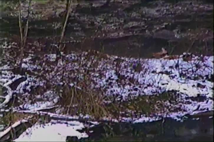 A vintage tilt-down shot reveals a snowy forest, a rocky cliff, and a cold river with ice floating on the surface