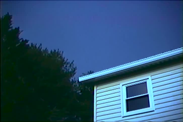 A powerful lightning strike illuminates the dark, stormy sky over a suburban house at night, captured on vintage footage