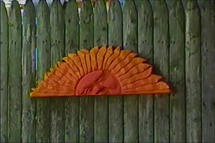 A vintage, static shot of a carved wooden sun decoration with a face mounted on a rustic green fence