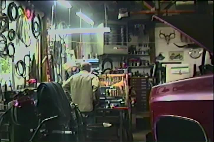 An elderly man works at his workbench in a classic, cluttered American garage next to his red pickup truck