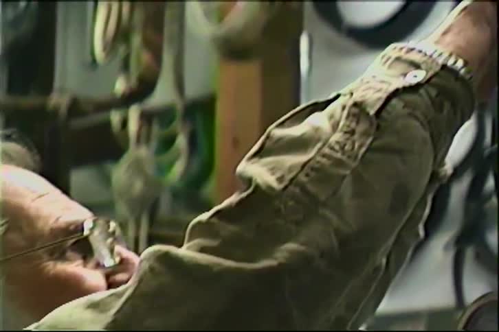 An elderly man with glasses carefully uses a wrench to work on a piece of machinery in his workshop