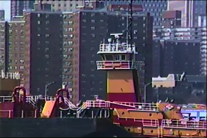 A colorful tugboat floats in an urban harbor with large city buildings in the background, captured on vintage VHS