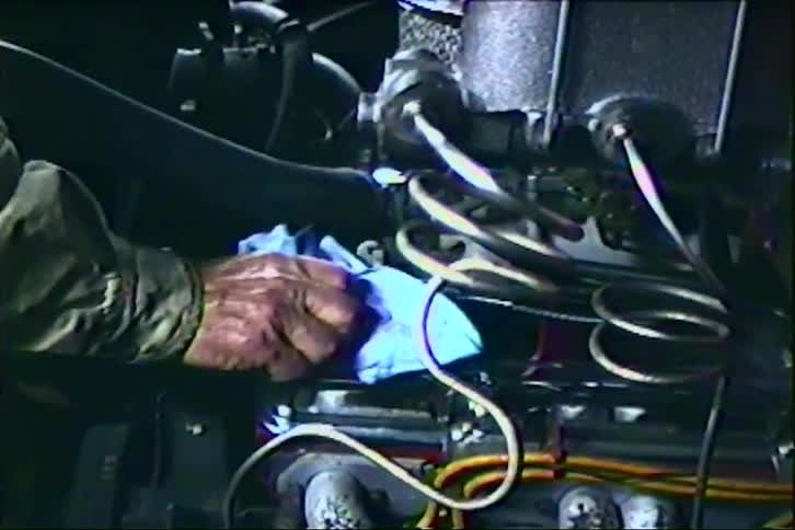 A mechanic with glasses carefully cleans a car engine with a blue cloth in his cluttered garage