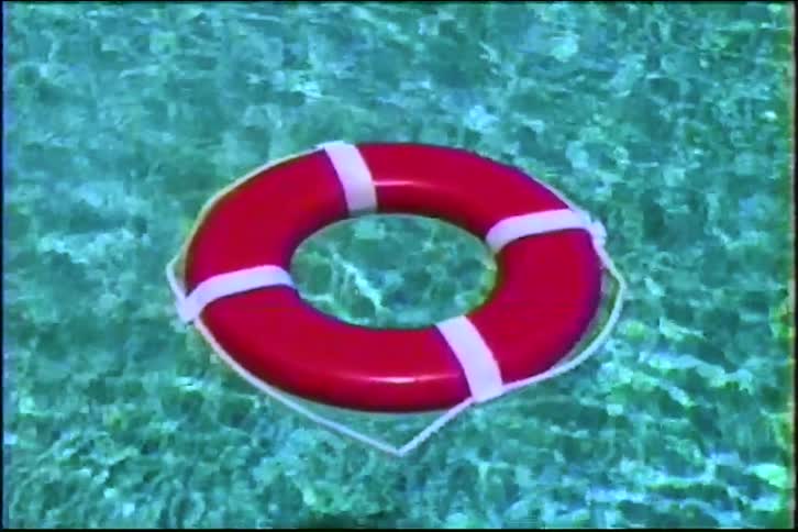 A red lifebuoy gently floats in the clear, sunlit turquoise water of a swimming pool in this vintage footage