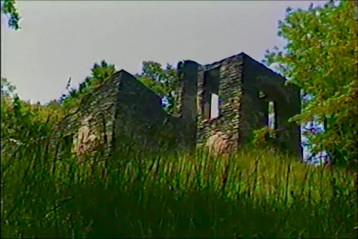 Vintage footage slowly zooms out from a low angle to reveal the crumbling stone walls of an abandoned building in the woods