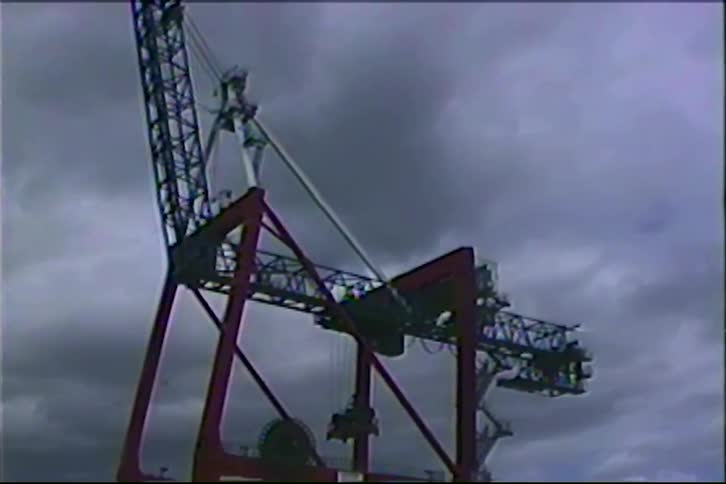 A low-angle shot tilts up a massive red industrial crane against a dark, cloudy sky