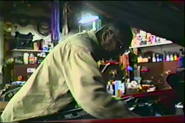 Vintage home video footage shows an older mechanic working on the engine of a red car in his cluttered garage