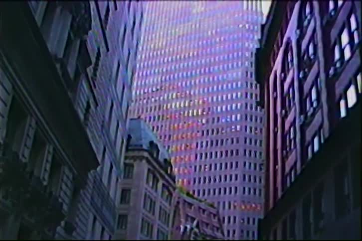 Vintage VHS footage captures a low-angle view of a massive modern skyscraper towering over a narrow, shadowed city street
