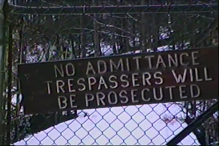 Vintage VHS footage slowly zooms in on a weathered 'No Admittance Trespassers Will Be Prosecuted' sign on a chain-link fence during winter
