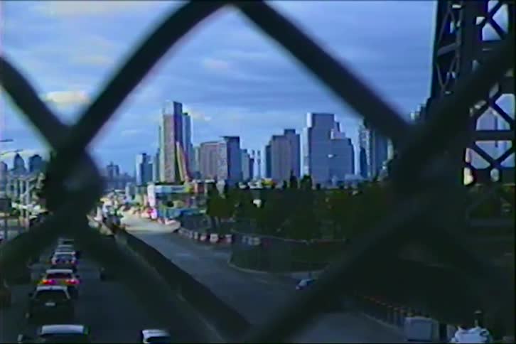 A vintage VHS-style zoom-in shot of a city skyline with traffic on a highway below, viewed through a chain-link fence