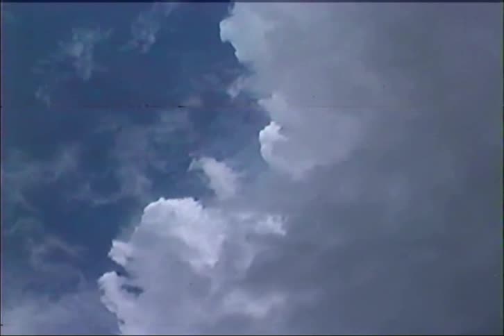 A vintage, low-angle, static shot of large fluffy clouds moving slowly across a deep blue sky