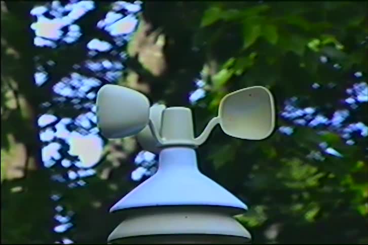 A vintage close-up shot of a weather station anemometer spinning slowly with out-of-focus green trees in the background