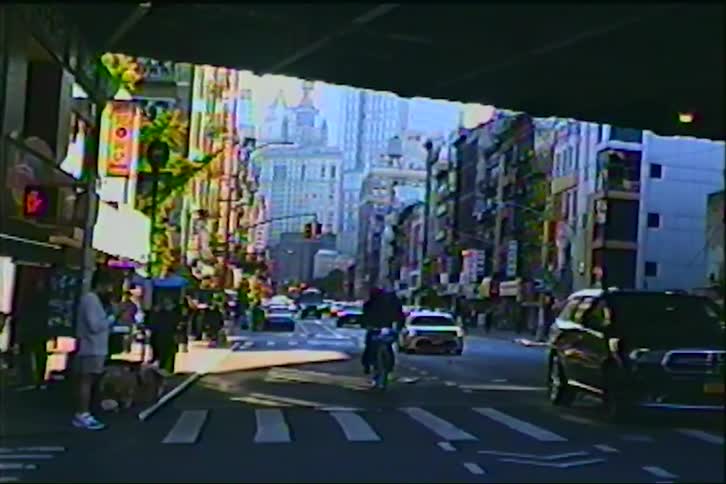 A retro VHS-style shot captures a busy New York City street scene with traffic and pedestrians from under an overpass