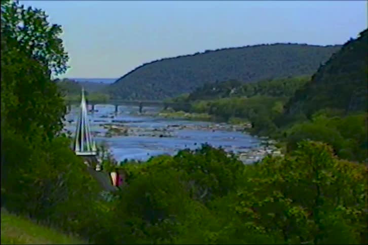 An archival shot slowly zooms in on a picturesque river flowing through a lush green valley, with a church steeple visible in the foreground