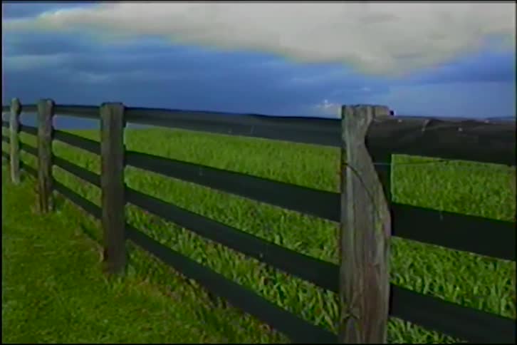 A rustic wooden fence lines a vibrant green pasture under a dramatic, stormy sky