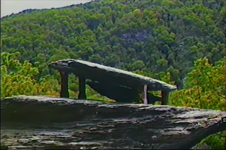 Vintage VHS footage slowly zooms in on a dolmen-like rock formation perched on a cliff overlooking a dense, green forest