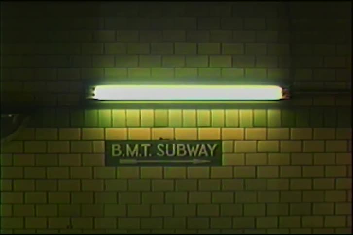 A flickering fluorescent light illuminates a vintage B.M.T. Subway sign on a tiled wall in a New York City station
