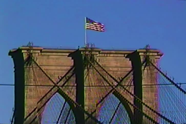 A vintage VHS-style shot of the American flag waving atop the iconic Brooklyn Bridge as the camera slowly zooms out