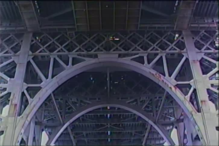 A vintage, low-angle shot slowly pans down the intricate steel underbelly of a massive bridge, revealing the World Trade Center towers framed by its iconic arch