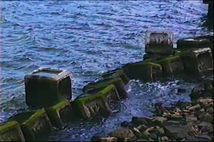 A nostalgic VHS-style recording shows gentle sea waves lapping against a moss-covered concrete block sea defense