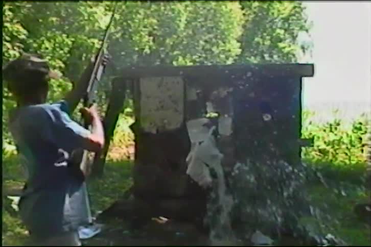 A young man shoots a rifle at a target in a field, hitting a water bottle in dramatic slow motion