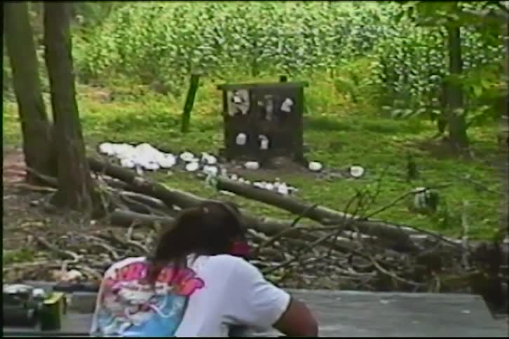 A person at a makeshift shooting range in the woods shoots a rifle at a target, causing a plastic jug to explode