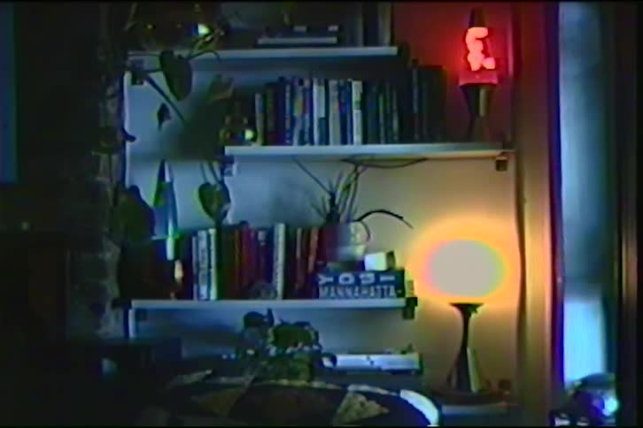 Vintage VHS footage shows a dimly lit room with a bookshelf, a warm glowing lamp, and a classic red lava lamp