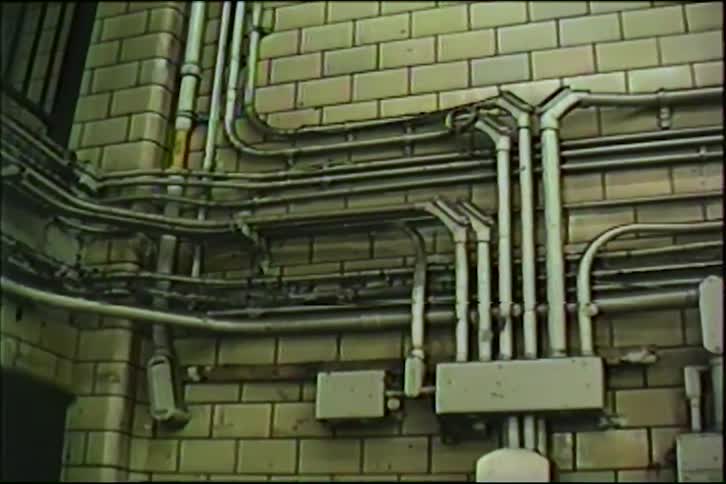 Vintage footage pans across a complex network of industrial electrical conduits and junction boxes on a tiled wall