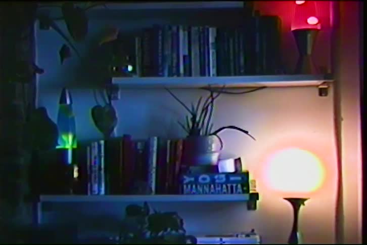A static shot of a dimly lit bookshelf with two lava lamps and an orb light, captured on grainy VHS tape