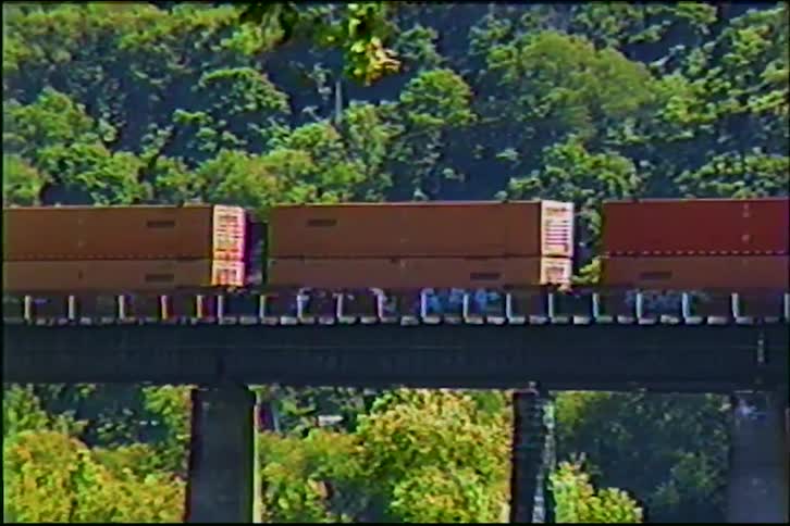 A freight train with shipping containers and graffiti slowly crosses a railway bridge over a river surrounded by lush green trees