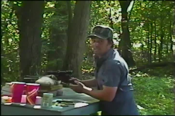 Vintage VHS footage shows a man target practicing with a scoped pistol in the woods, smiling proudly after his shot