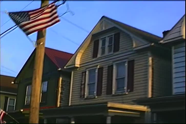 A retro VHS-style pan across a row of colorful American suburban homes on a quiet street in the evening