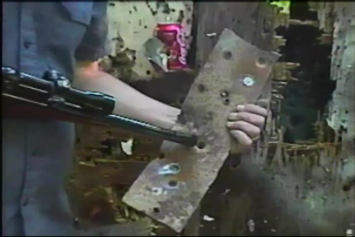 A man holds up a metal plate with bullet holes and compares the size of one hole to the barrel of his rifle in vintage VHS footage