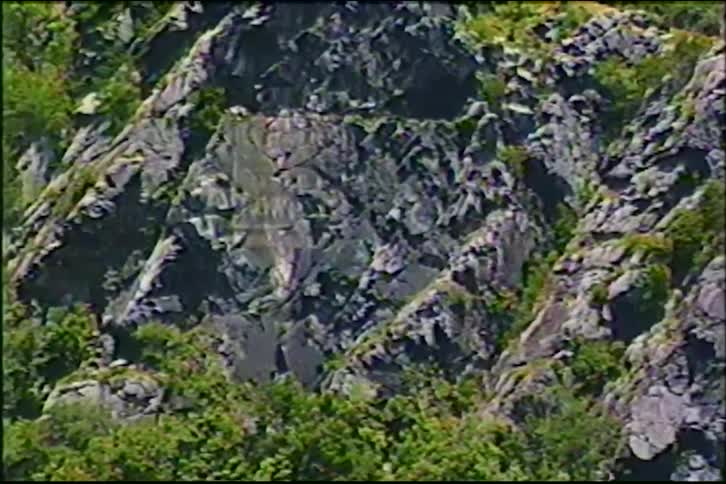 Vintage footage zooms in on a rocky, tree-covered mountain with a railway bridge and tunnel crossing a river below