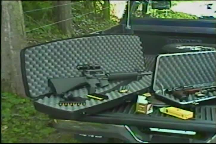 A collection of rifles and ammunition are displayed in open cases on the tailgate of a pickup truck in a wooded area