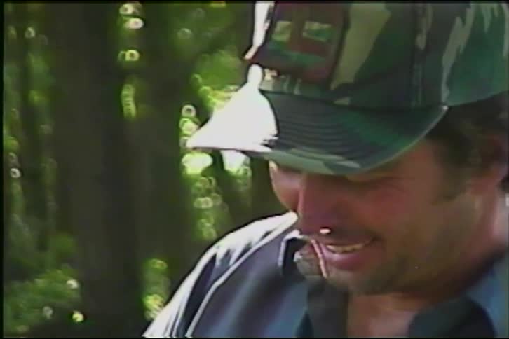 A smiling man in a camo hat laughs heartily while at a makeshift shooting range in a sunny, green forest