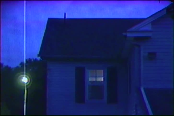 Vintage VHS footage shows a static shot of a suburban house at dusk with a single illuminated window before cutting to static