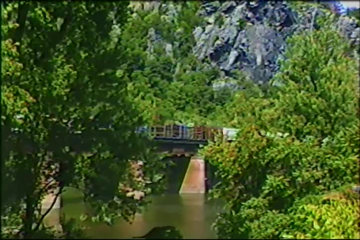 A vintage shot of a long freight train slowly crossing a bridge over a river in a lush, green mountain landscape